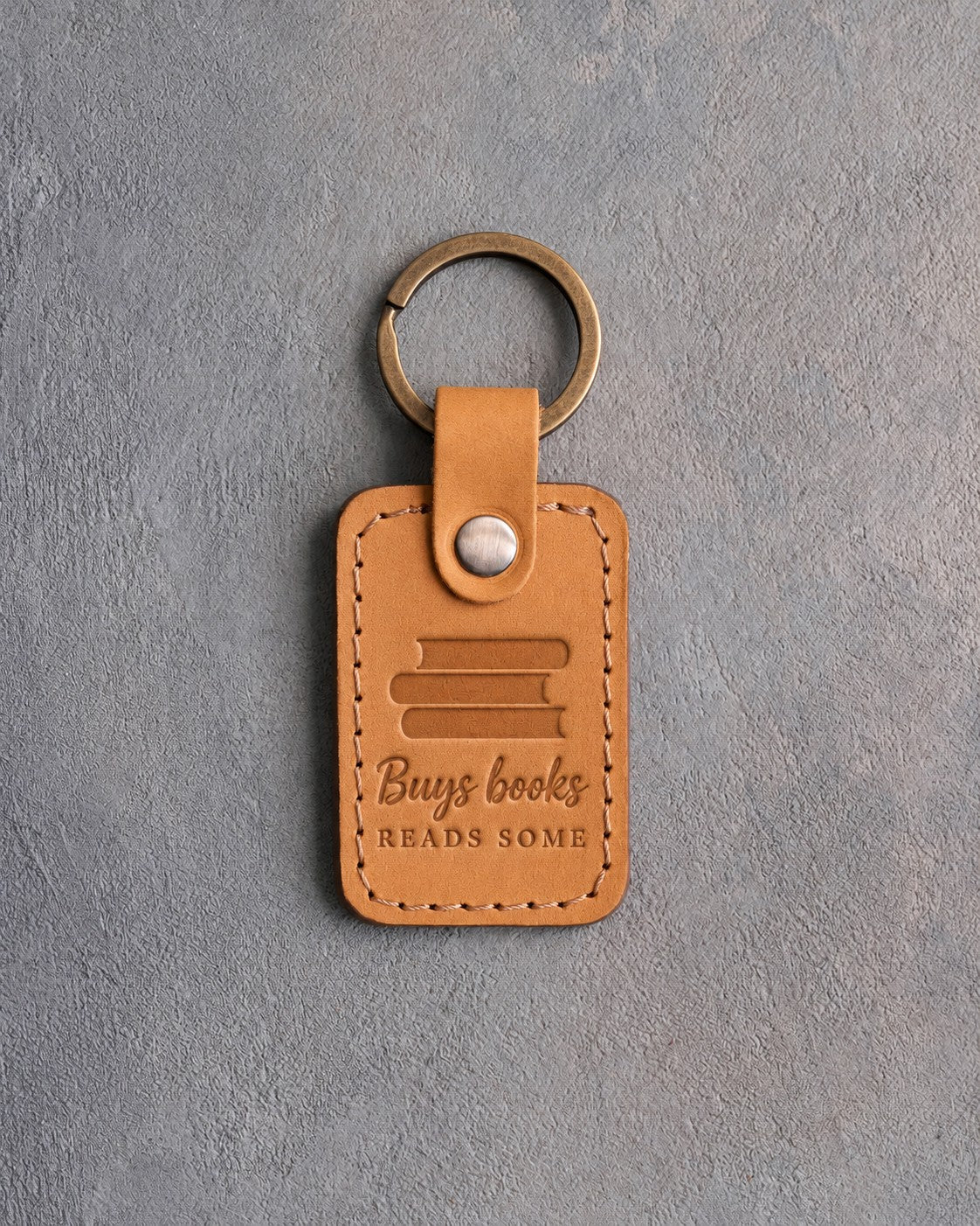 Indian Summer tan leather keychain debossed with a pile of books above Buys books and READS SOME, on gray concrete