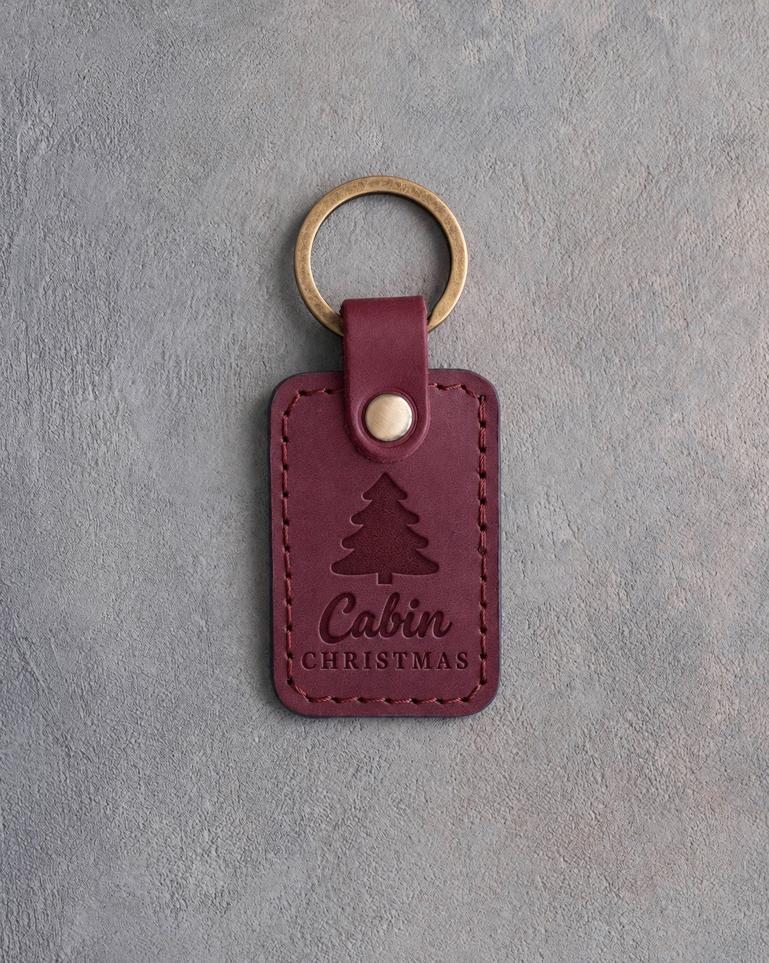 Sangria wine red leather keychain debossed with a single spruce tree above the words Cabin CHRISTMAS, on gray plaster
