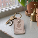 Gray sand Cactus Collector keychain on a shelf lined with small labeled pots of cacti