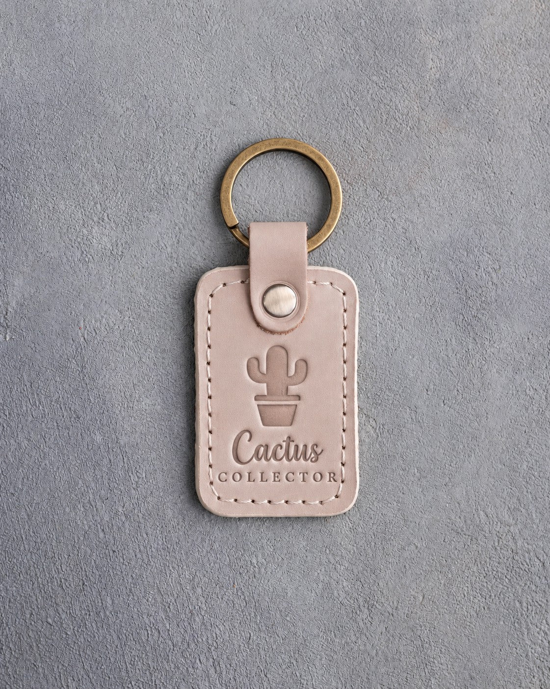 Gray sand leather keychain debossed with a potted cactus above Cactus COLLECTOR, on gray concrete