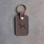 Country gray leather keychain debossed with a Cairn Terrier silhouette above THE CAIRN, on gray concrete