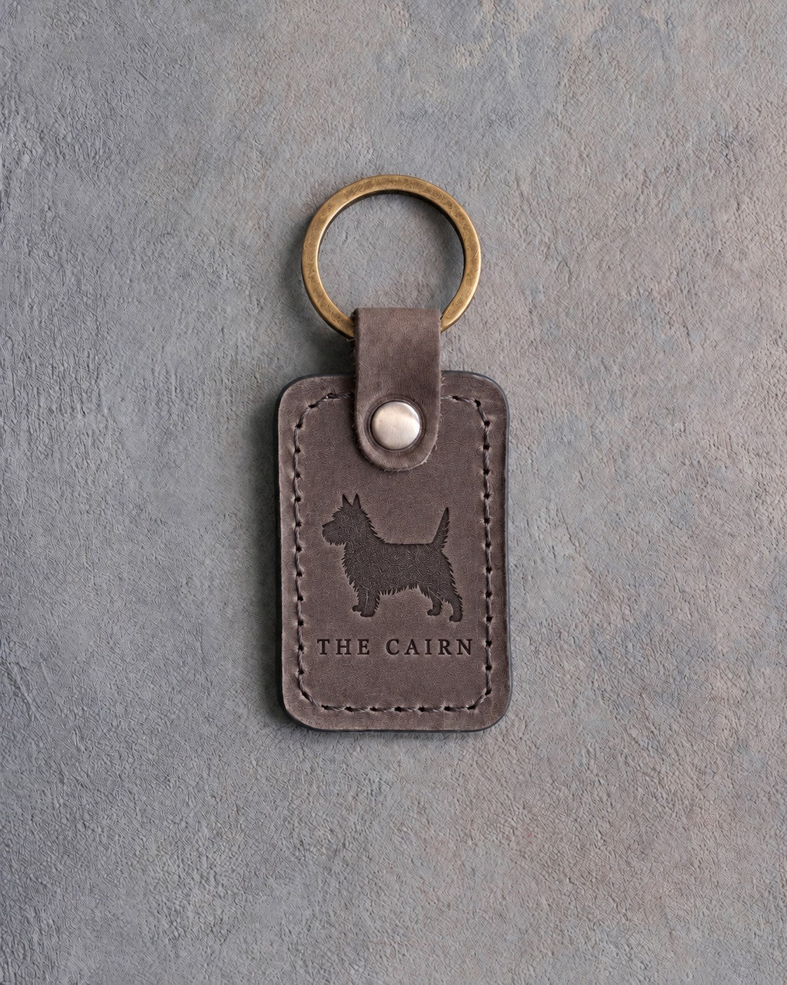 Country gray leather keychain debossed with a Cairn Terrier silhouette above THE CAIRN, on gray concrete