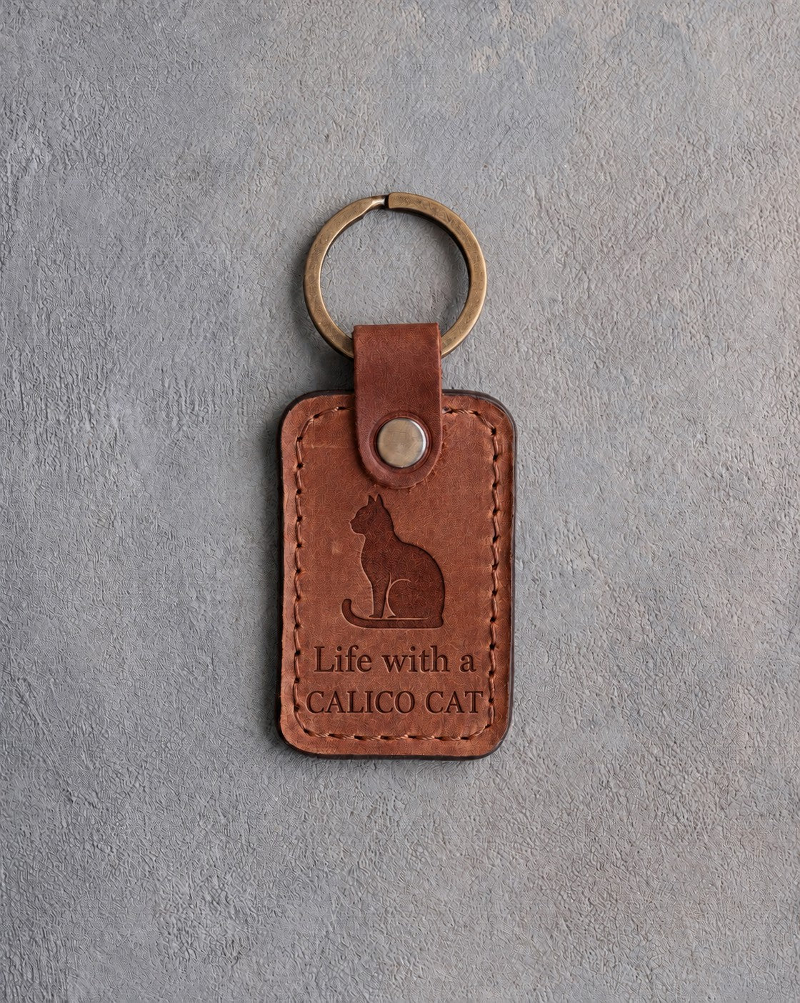 Arizona tan leather keychain debossed with a sitting cat above Life with a and CALICO CAT, on gray concrete