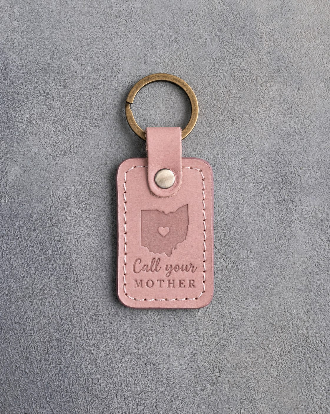 Blush pink leather keychain debossed with an Ohio silhouette and a heart at Columbus above Call your MOTHER, on gray concrete