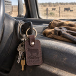 Dark espresso Calving SEASON keychain on the ignition key of a ranch pickup, work gloves on the dash and a pasture through the windshield