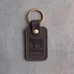 Dark espresso leather keychain debossed with a steer silhouette above Calving and SEASON, on gray plaster