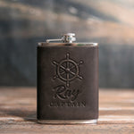 Dark espresso leather-wrapped hip flask debossed with a six-spoke ship's wheel above Ray and CAPTAIN, on a wooden table
