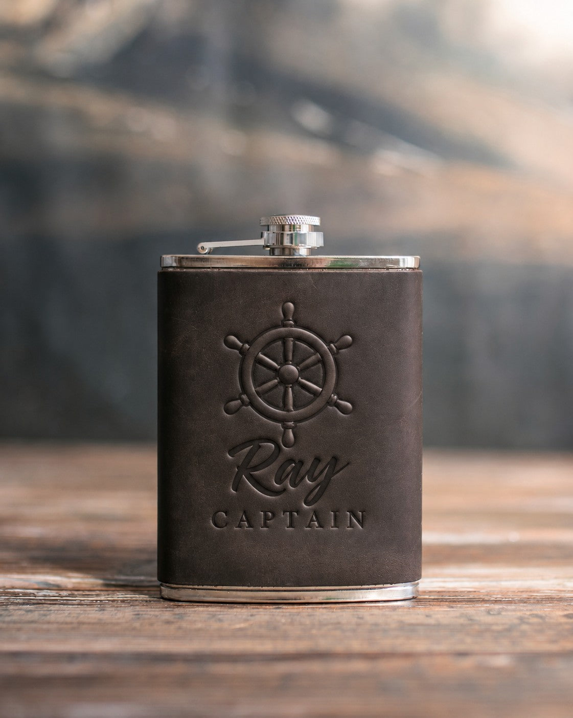 Dark espresso leather-wrapped hip flask debossed with a six-spoke ship's wheel above Ray and CAPTAIN, on a wooden table