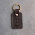Dark espresso leather keychain debossed with a six-spoke ship's wheel above Ray and CAPTAIN, on gray plaster