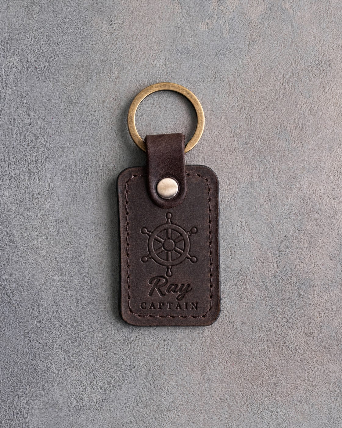 Dark espresso leather keychain debossed with a six-spoke ship's wheel above Ray and CAPTAIN, on gray plaster