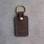 Dark espresso leather keychain debossed with a ship's wheel above CAPTAIN, on gray concrete