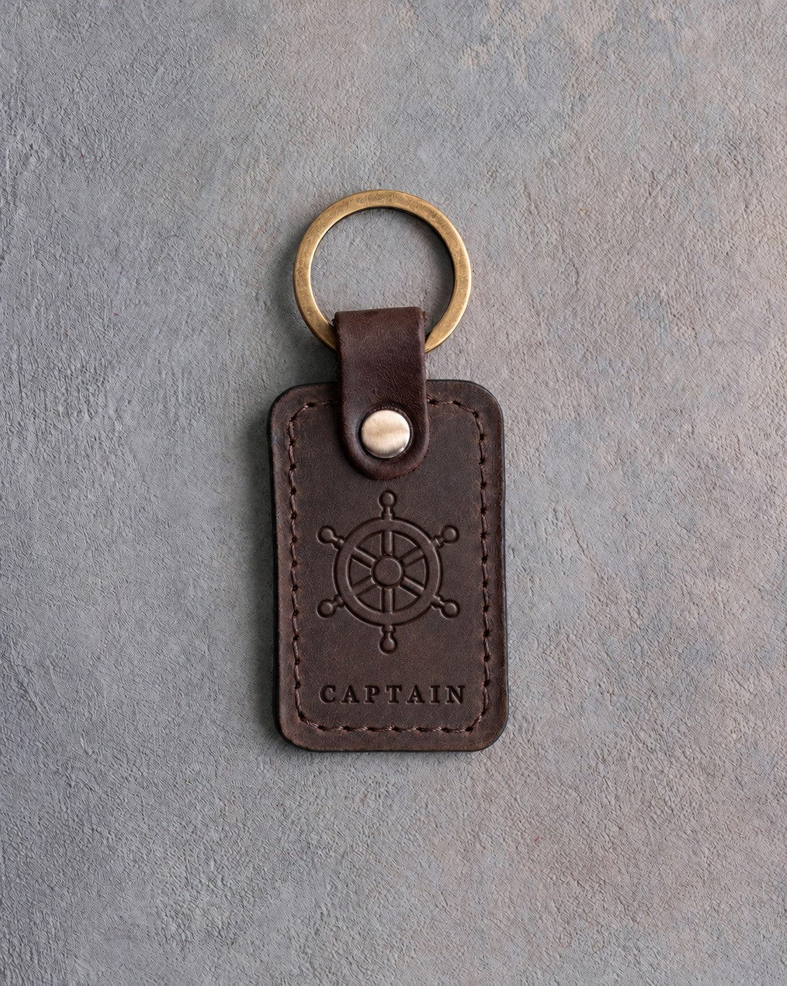 Dark espresso leather keychain debossed with a ship's wheel above CAPTAIN, on gray concrete