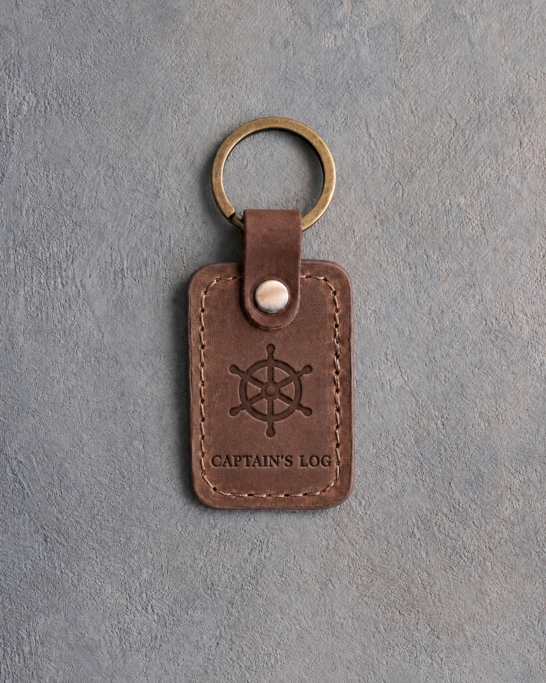 Sicilian Brown leather keychain debossed with a six-spoke ship's wheel above CAPTAIN'S LOG, on gray concrete