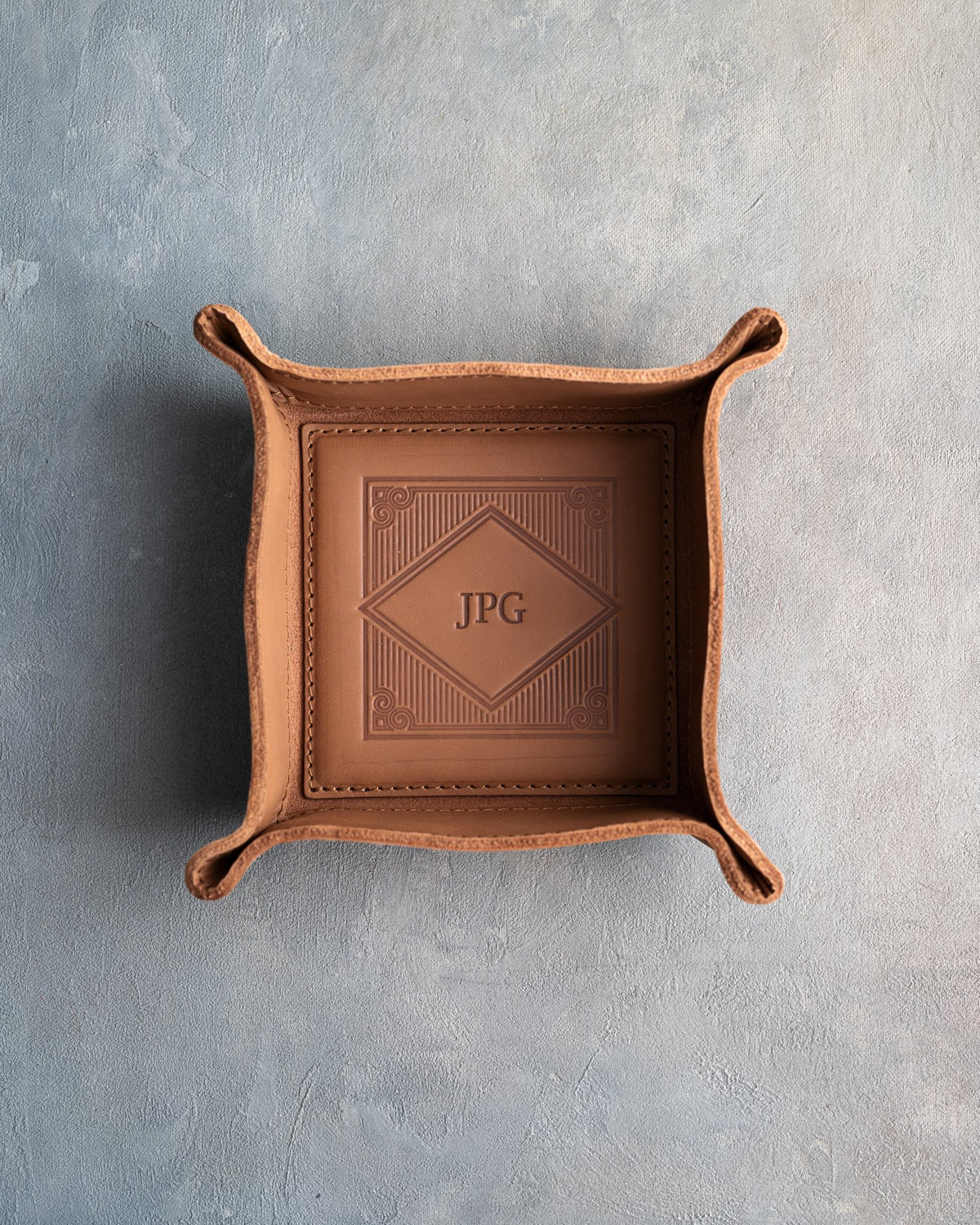 Caramel leather valet tray with snapped corners and the initials JPG pressed into the diamond frame, seen from above