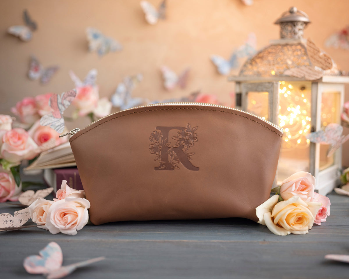 Caramel initial makeup pouch with a floral monogram, staged among pale roses on a slate table