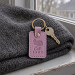 Lilac Cat Lady keychain beside a teacup and an open paperback on a cozy armchair