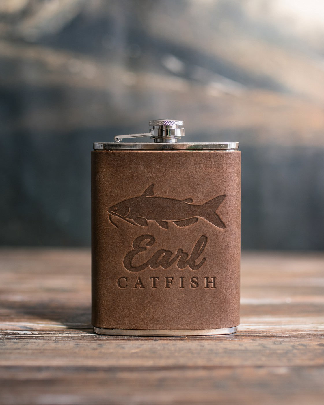 Sicilian brown leather-wrapped hip flask debossed with a channel catfish above Earl and CATFISH, on a wooden table