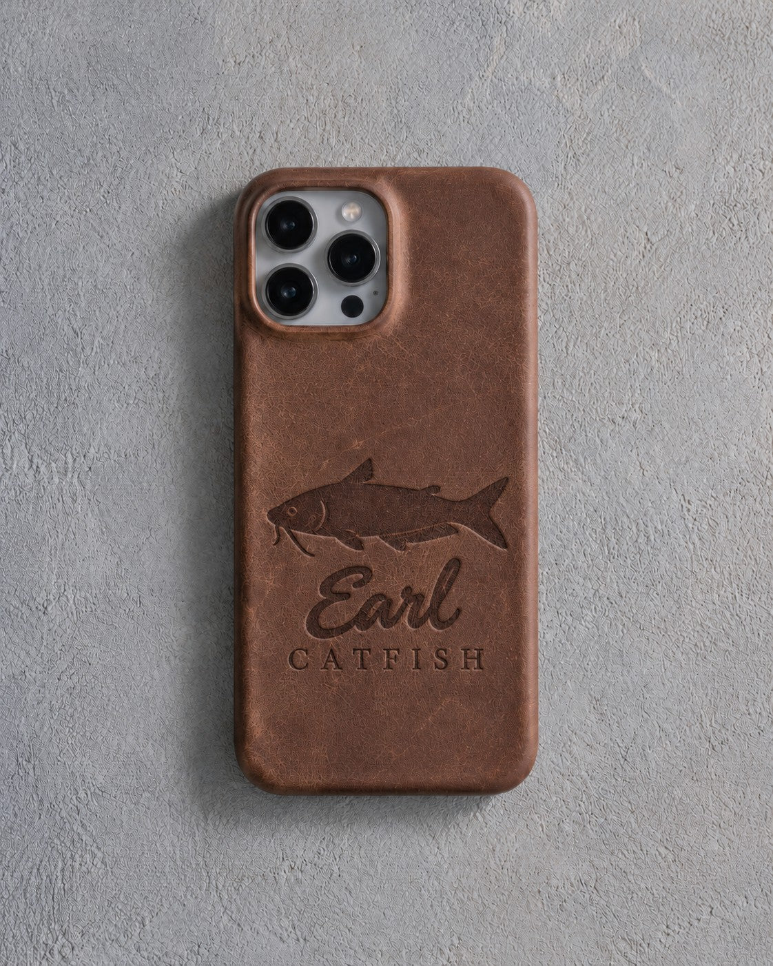 Back of a Sicilian brown leather iPhone case debossed with a channel catfish above Earl and CATFISH below the camera, on gray plaster