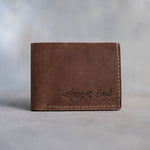 Closed Sicilian brown leather bifold wallet with a channel catfish beside Earl and CATFISH pressed in the lower right corner, on gray plaster
