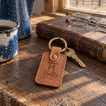 Hollis CATTLE CO keychain in caramel leather on a dark oak kitchen table next to reading glasses and a closed ranch ledger