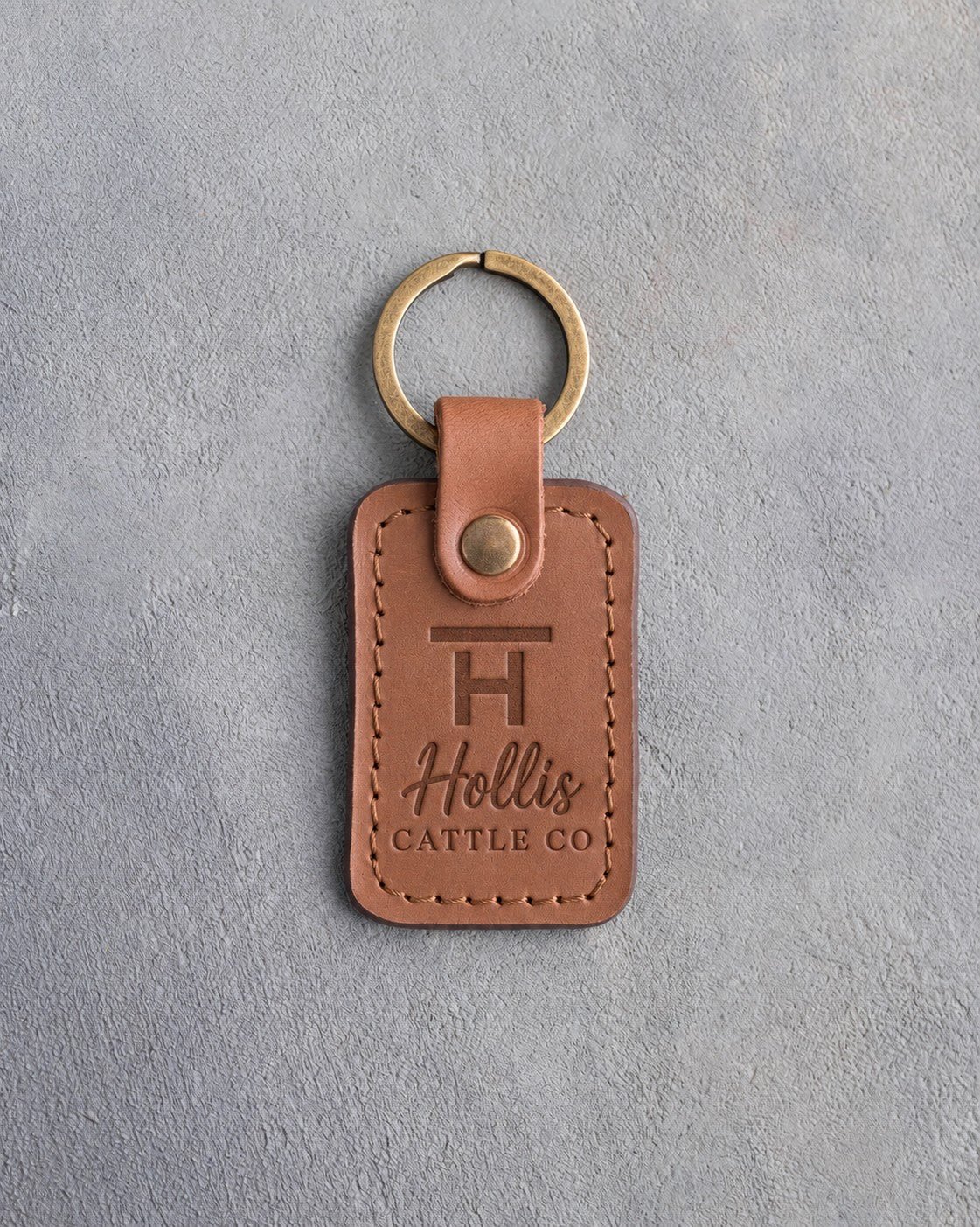 Caramel leather keychain debossed with a Bar H ranch brand above the name Hollis and CATTLE CO, on gray plaster