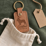 Caramel Certified Snorer keychain peeking from a cotton gift pouch on green linen next to a kraft tag