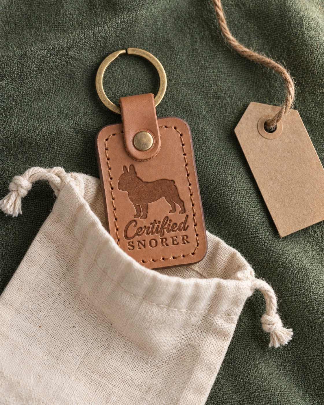 Caramel Certified Snorer keychain peeking from a cotton gift pouch on green linen next to a kraft tag