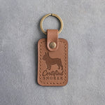 Caramel leather keychain with a debossed French bulldog and the words Certified SNORER on gray concrete
