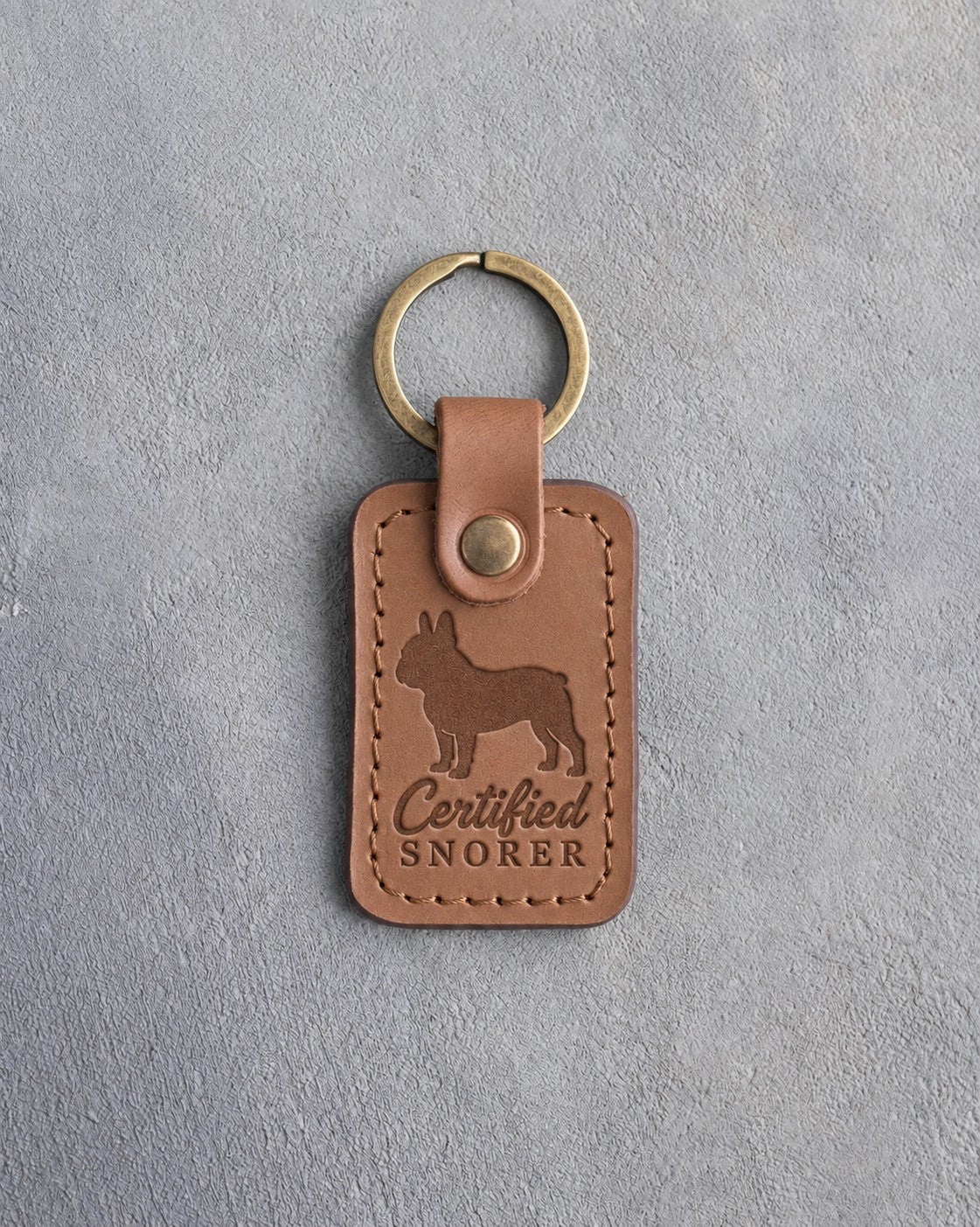 Caramel leather keychain with a debossed French bulldog and the words Certified SNORER on gray concrete