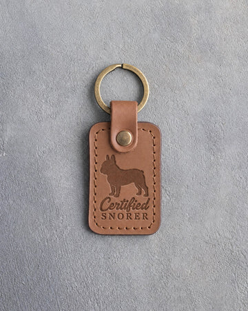 Caramel leather keychain with a debossed French bulldog and the words Certified SNORER on gray concrete