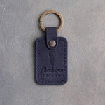 Blueberry navy leather keychain debossed with a handheld microphone above the words Check one CHECK TWO, on gray plaster
