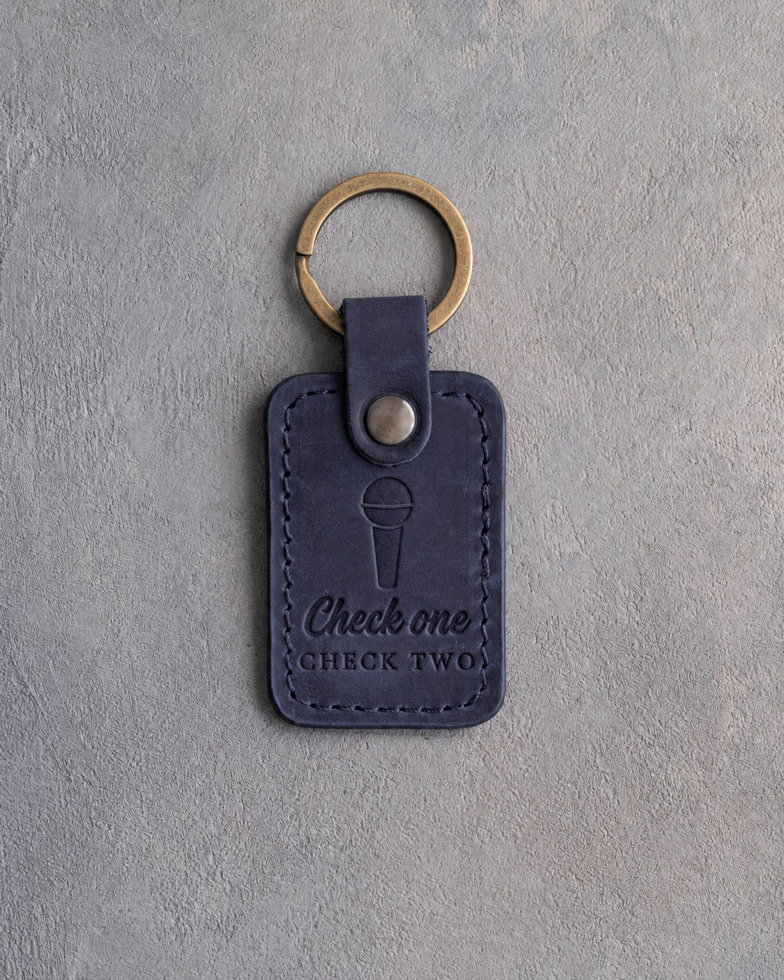 Blueberry navy leather keychain debossed with a handheld microphone above the words Check one CHECK TWO, on gray plaster