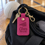 Bright pink Cheer Mom keychain lying on an open black duffel bag on a hardwood gym floor