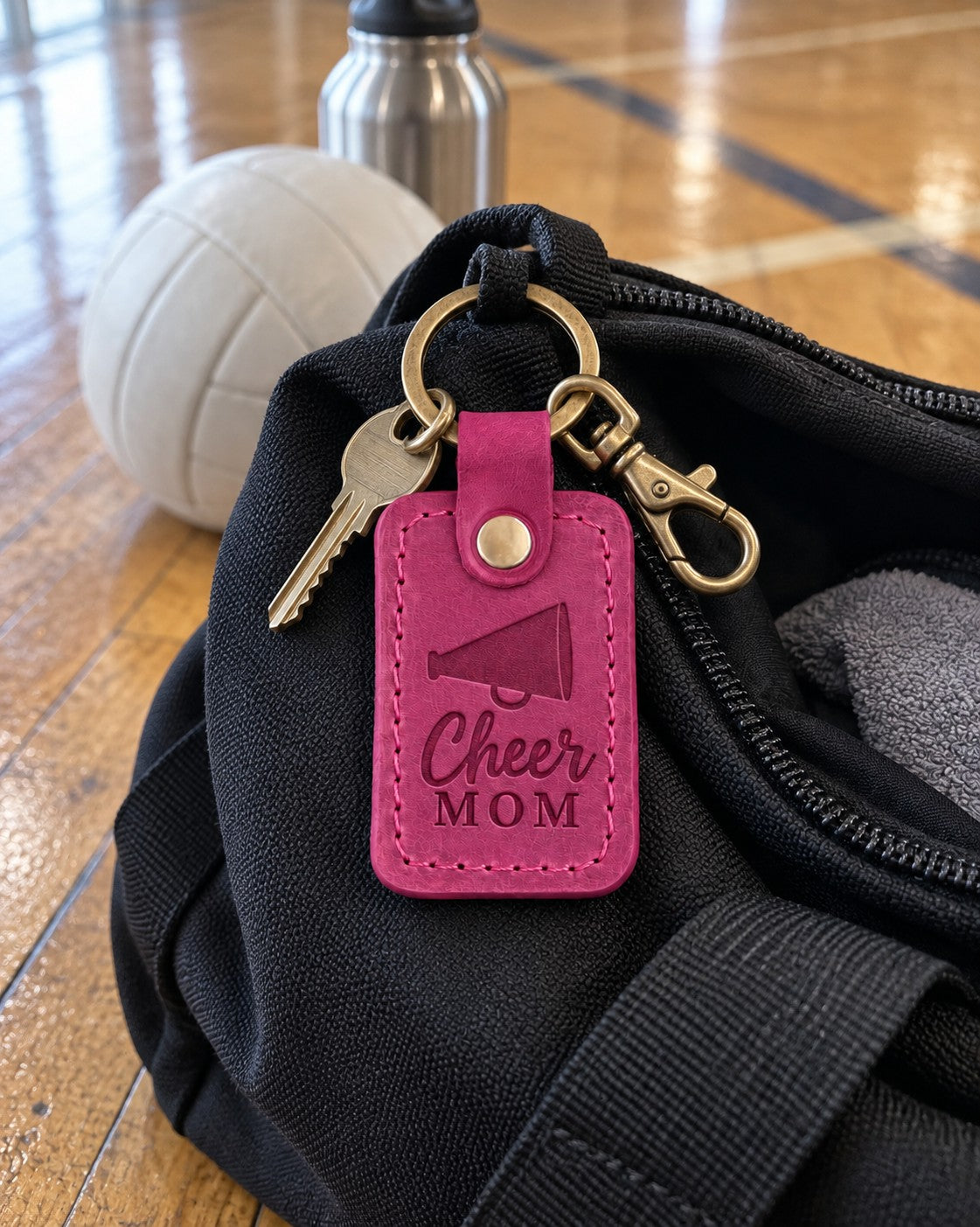 Bright pink Cheer Mom keychain lying on an open black duffel bag on a hardwood gym floor
