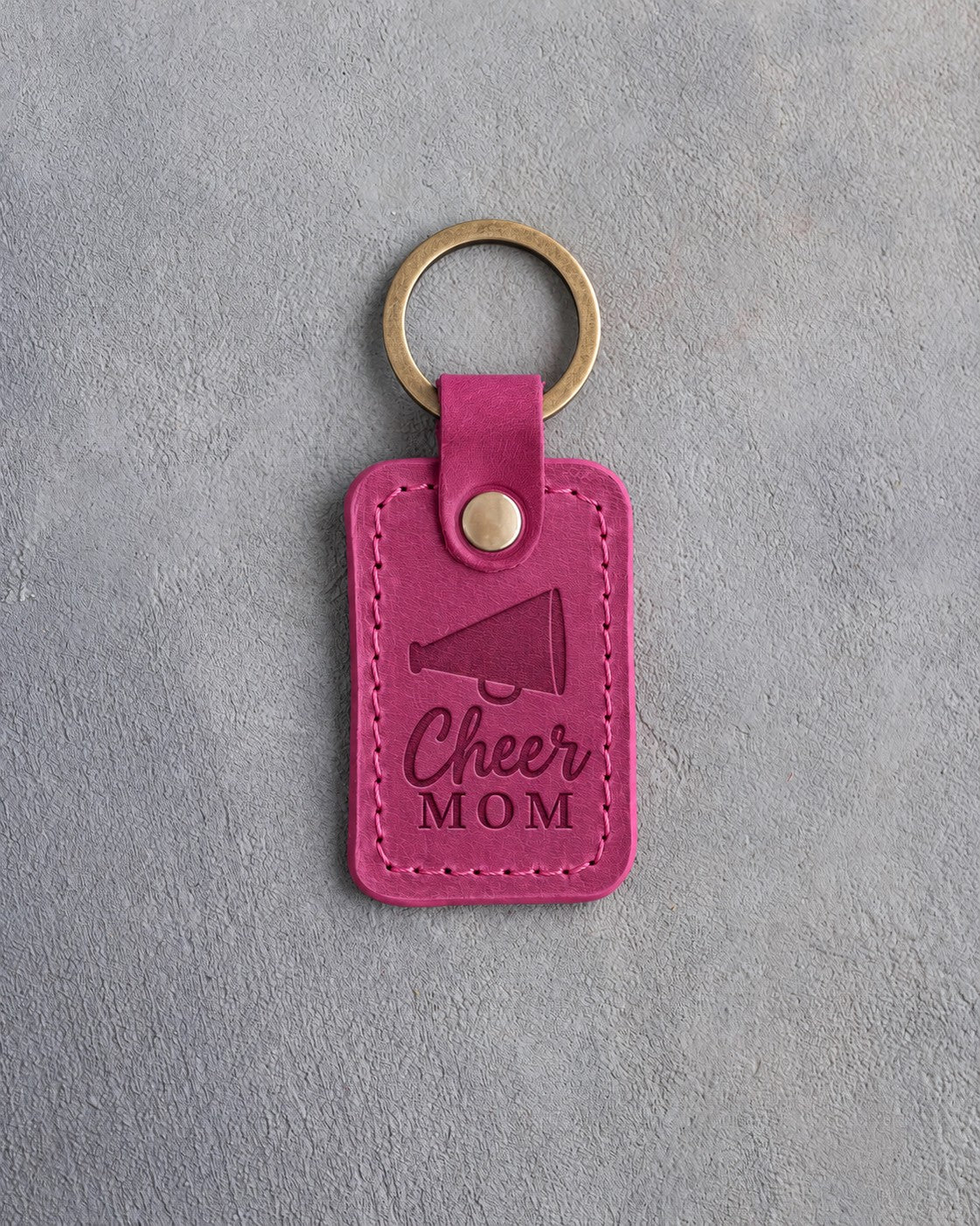 Fuchsia pink leather keychain debossed with a megaphone and the words Cheer MOM, on gray plaster