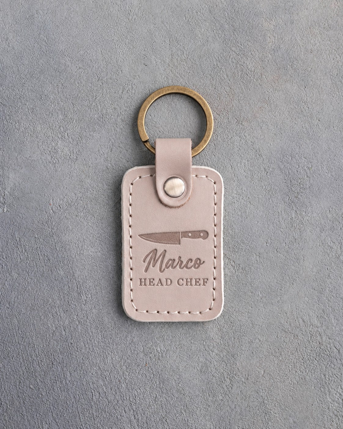 Gray sand leather keychain debossed with a chef's knife above the name Marco and HEAD CHEF, on gray plaster