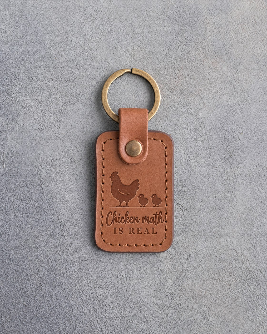 Caramel leather keychain debossed with a hen and two chicks above the words Chicken math IS REAL, on gray concrete