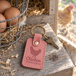 Rose pink Chicken Whisperer keychain on a weathered pine nesting box ledge beside brown eggs in a wire basket, a hen soft in the run behind