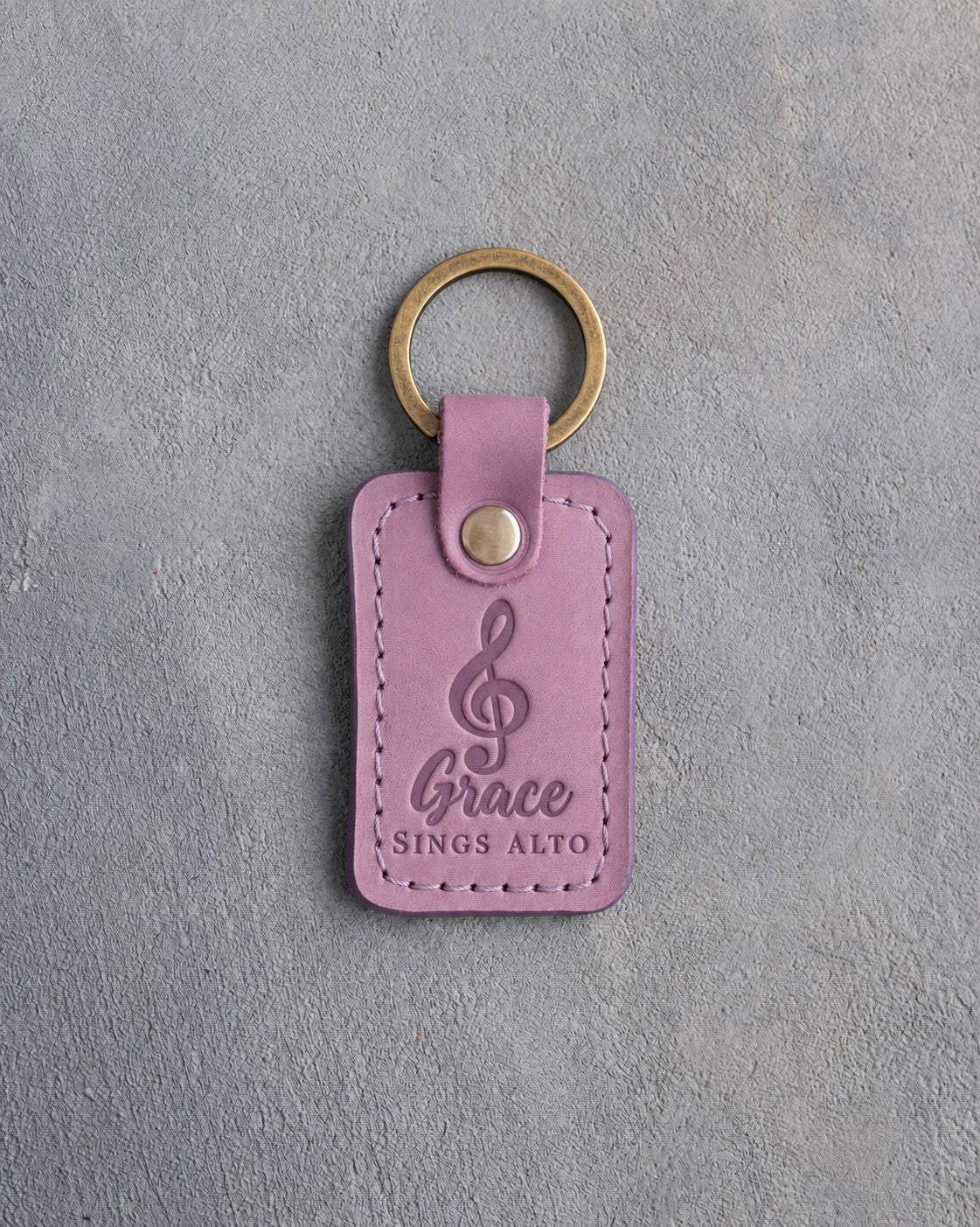 Lilac leather keychain debossed with a treble clef above the name Grace and SINGS ALTO, on gray plaster