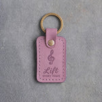 Lilac leather keychain debossed with a treble clef above the words Lift and EVERY VOICE, on gray concrete