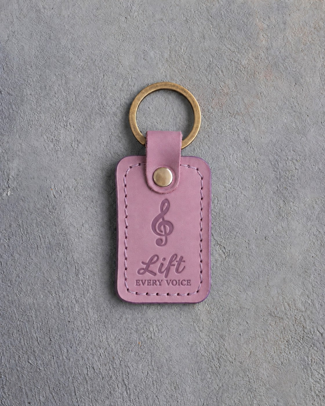 Lilac leather keychain debossed with a treble clef above the words Lift and EVERY VOICE, on gray concrete