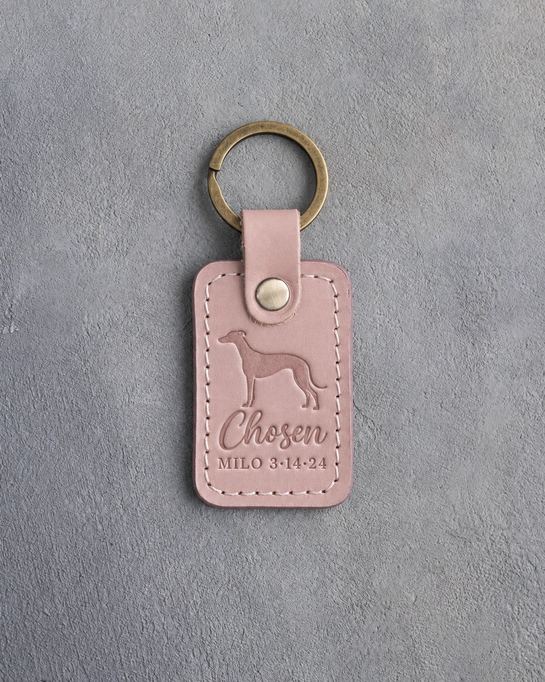 Blush pink leather keychain debossed with a greyhound above Chosen and MILO 3·14·24, on gray concrete