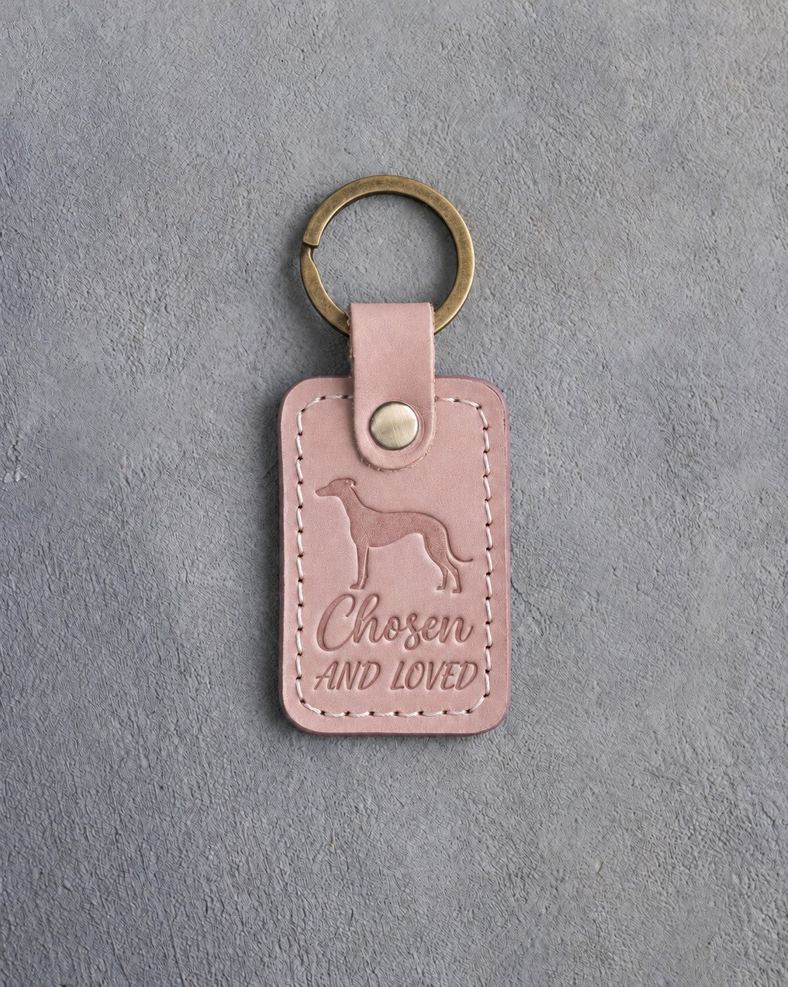 Blush pink leather keychain debossed with a greyhound above Chosen and AND LOVED, on gray concrete