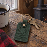 Christmas at Deer Camp keychain in forest green leather hanging from a cabin key beside a woodstove with a stocking above it