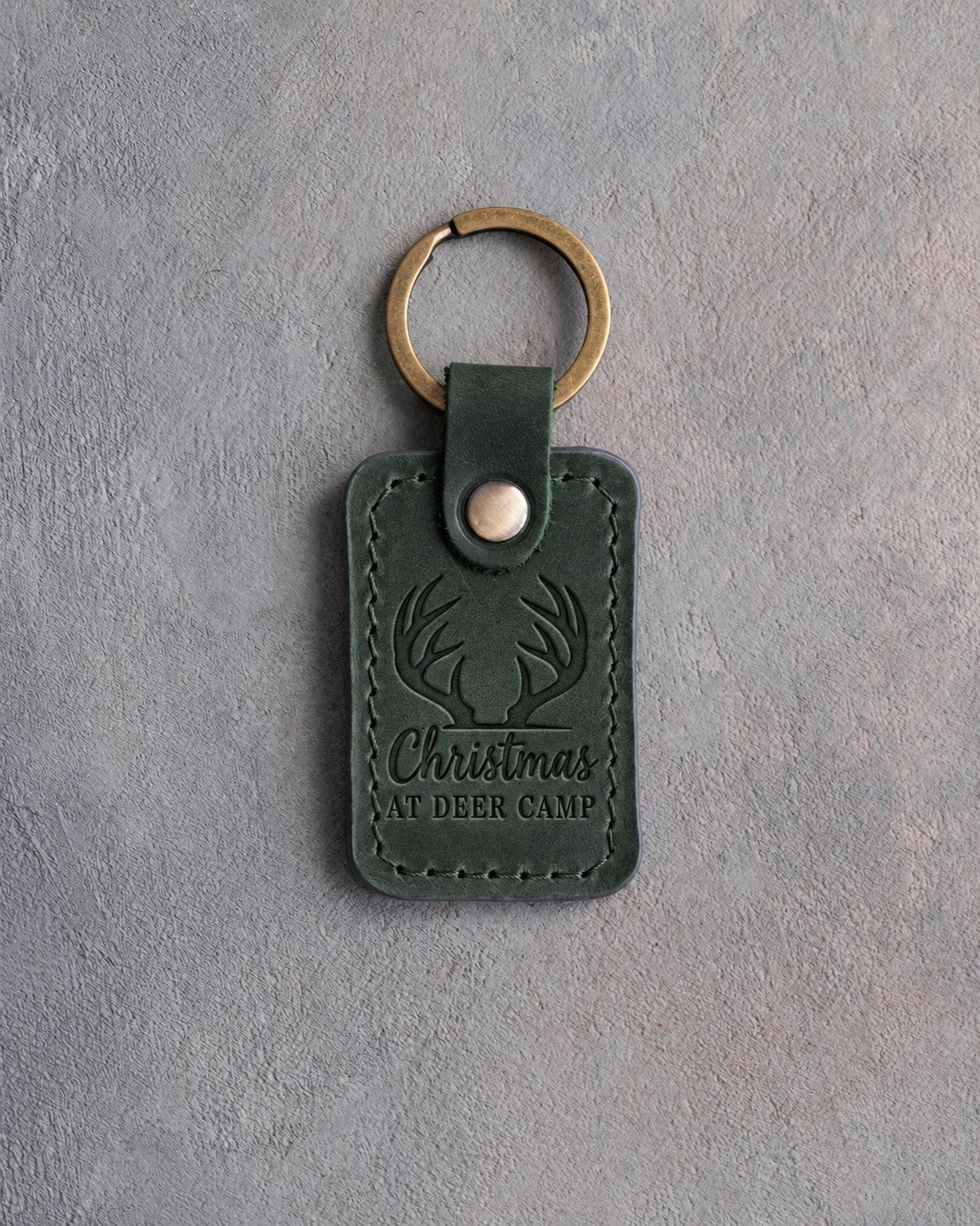 Forest green leather keychain debossed with a front view of whitetail antlers above the words Christmas AT DEER CAMP, on gray plaster