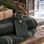 Forest green Christmas AT THE RANCH keychain on a folded green wool blanket on a pine bench inside a barn door, snow on the corral rails beyond