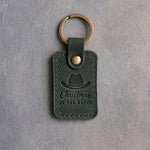 Forest green leather keychain debossed with a cowboy hat above Christmas and AT THE RANCH, on gray plaster
