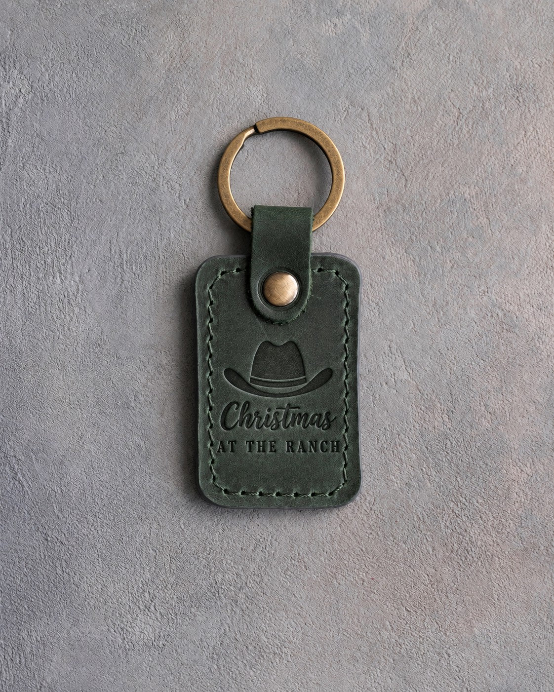 Forest green leather keychain debossed with a cowboy hat above Christmas and AT THE RANCH, on gray plaster