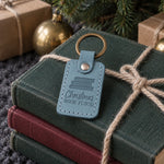 Turquoise Christmas BOOK FLOOD keychain on a yarn-tied stack of books beneath a fir tree