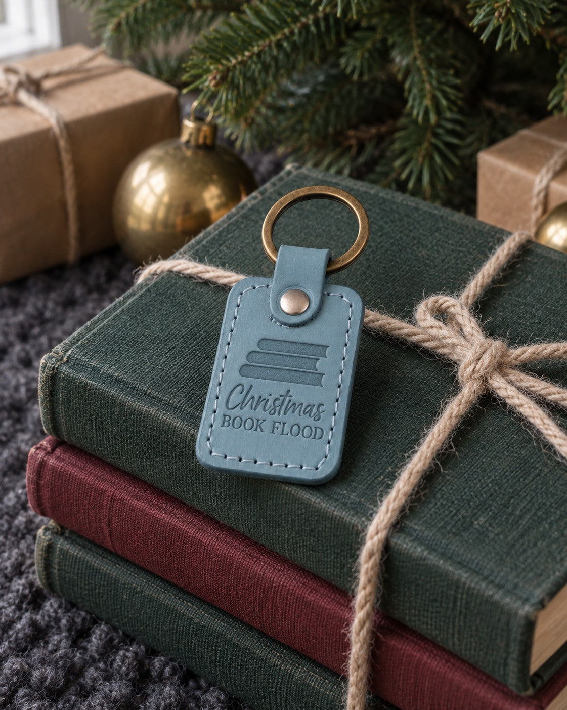 Turquoise Christmas BOOK FLOOD keychain on a yarn-tied stack of books beneath a fir tree